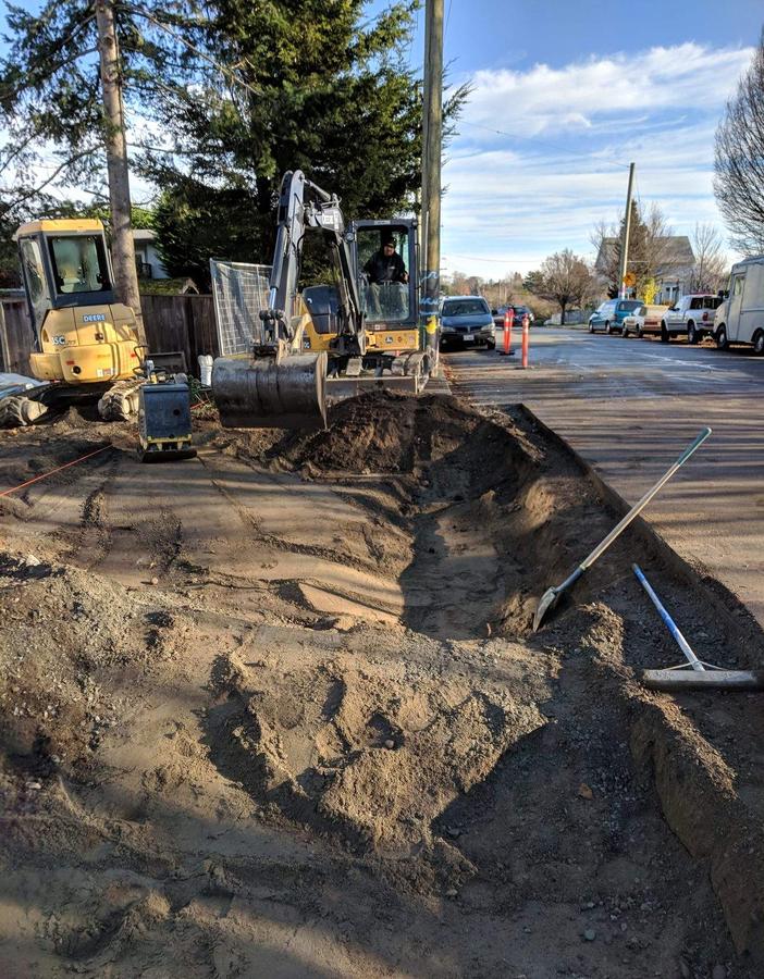 Mini excavator digging a service trench on a Greater Victoria street, operator in the cab